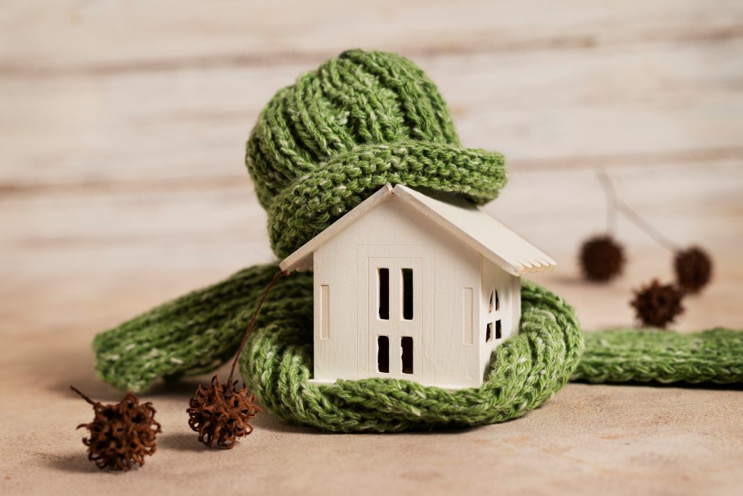 Small model house wrapped in green knitted scarf and hat, showing property care and home protection.